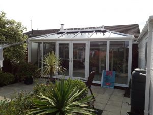 A traditional Edwardian conservatory in white in a back garden with palms in pots and a water butt on the right hand side.